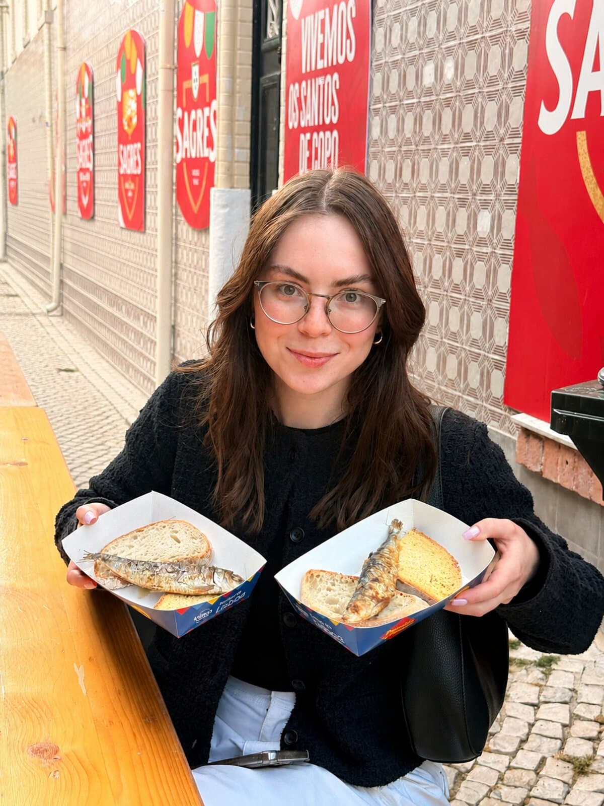 Karolina smiling with two trays of grilled sardines at the Santos festival in Lisbon